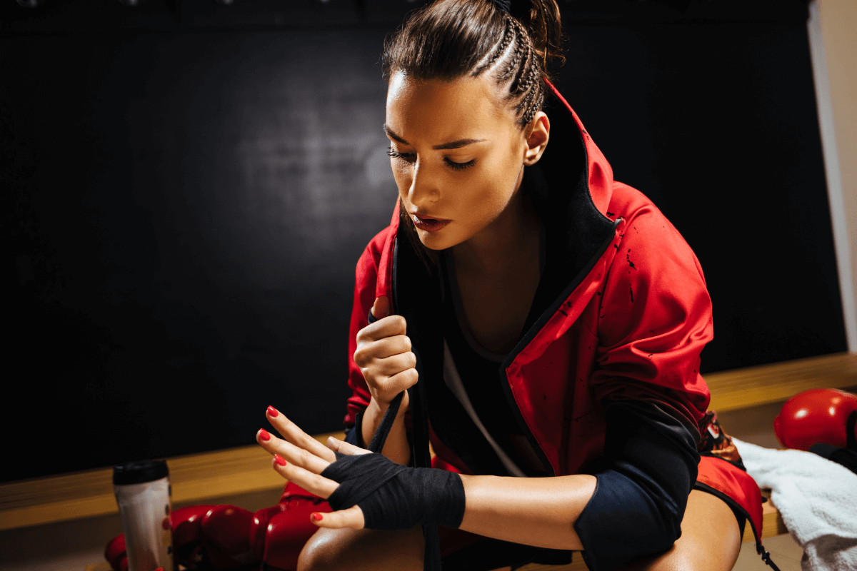 Lutadora sentada no banco ajustando a bandagem nas mãos, etapa fundamental para garantir a durabilidade do kit de Muay Thai e evitar lesões.
